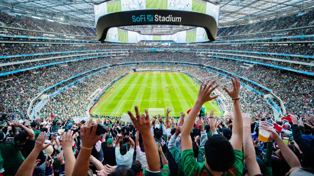 A vibrant panoramic view of SoFi Stadium during a packed football match, with cheering fans in the foreground and a clear view of the field and giant circular scoreboard.