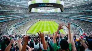 A vibrant panoramic view of SoFi Stadium during a packed football match, with cheering fans in the foreground and a clear view of the field and giant circular scoreboard.