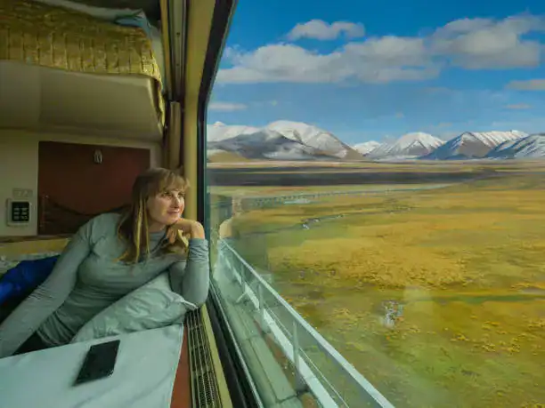 A woman looking out of a train window at a vast landscape of golden fields and snow-capped mountains under a blue sky.
