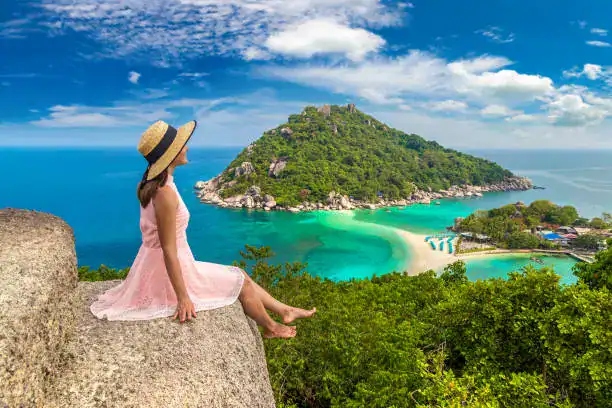 A woman in a pink dress and sun hat overlooks the turquoise waters and sandbar of Koh Nang Yuan, Thailand.