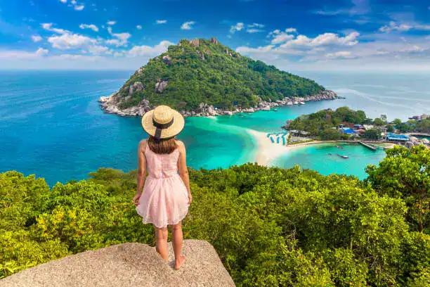 A woman in a pink dress and sun hat overlooks the turquoise waters and sandbar of Koh Nang Yuan, Thailand.