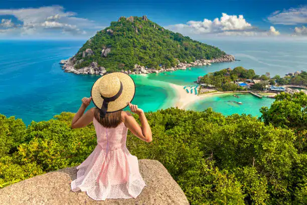 A woman in a pink dress and sun hat overlooks the turquoise waters and sandbar of Koh Nang Yuan, Thailand.