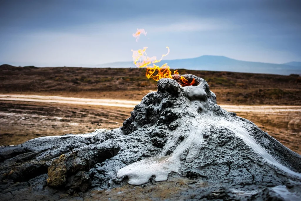 A natural mud volcano landscape near Baku, Azerbaijan, with small flames erupting from the top of a grey, bubbling mud mound.