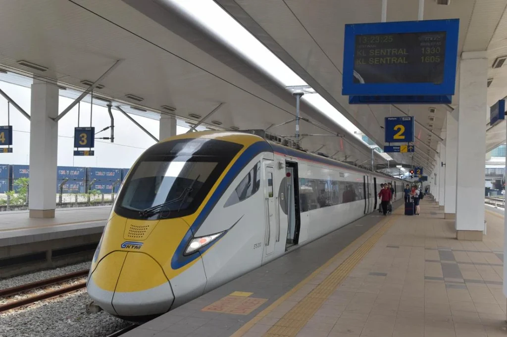 A high-speed KTM ETS electric train with a yellow and blue nose parked at a concrete station platform with passengers waiting in the background.