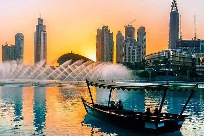 A traditional wooden abra boat carrying silhouettes of passengers on the Burj Lake in Dubai, with the dancing fountain and city skyline at sunset.