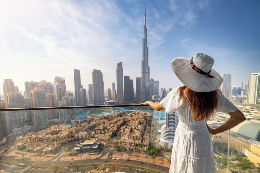 A woman in a sun hat and white dress enjoying a panoramic view of the Dubai skyline and the Burj Khalifa from a high-rise balcony.