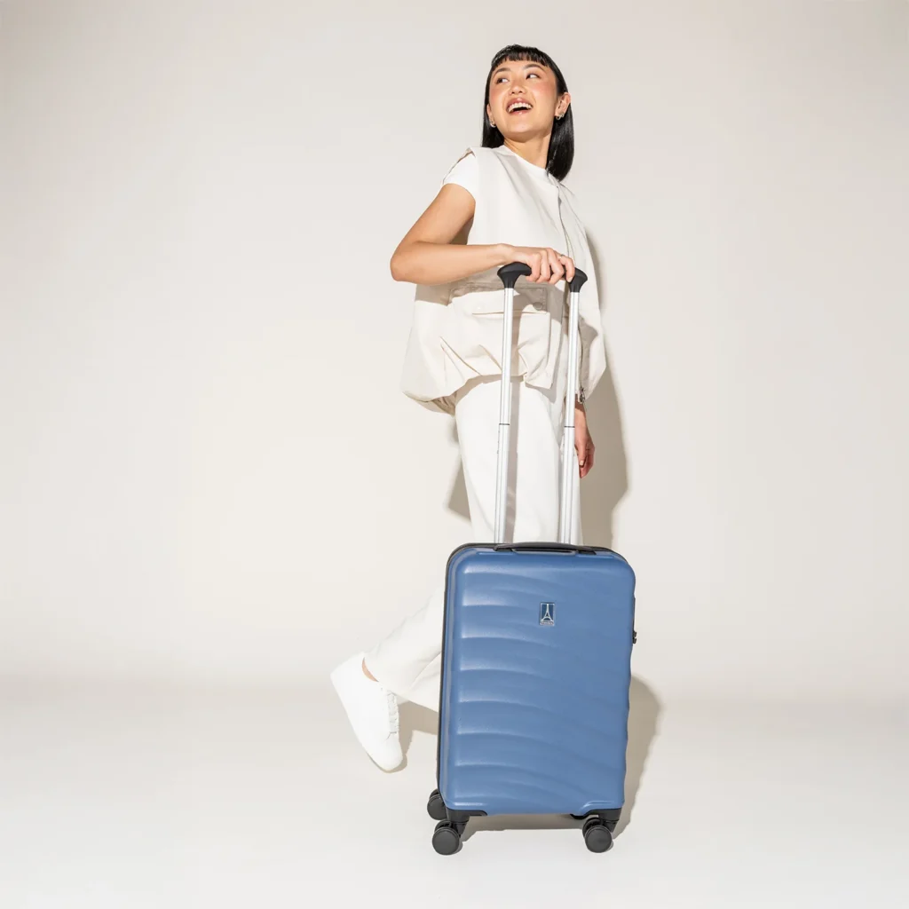 A smiling woman in an all-white outfit walking with a blue hard-shell carry-on suitcase against a plain white background.