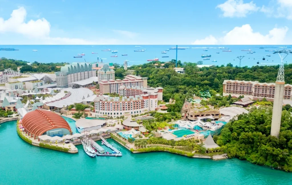 An aerial view of Resorts World Sentosa in Singapore, featuring hotels, water parks, a cable car tower, and a vibrant harbor with turquoise water.