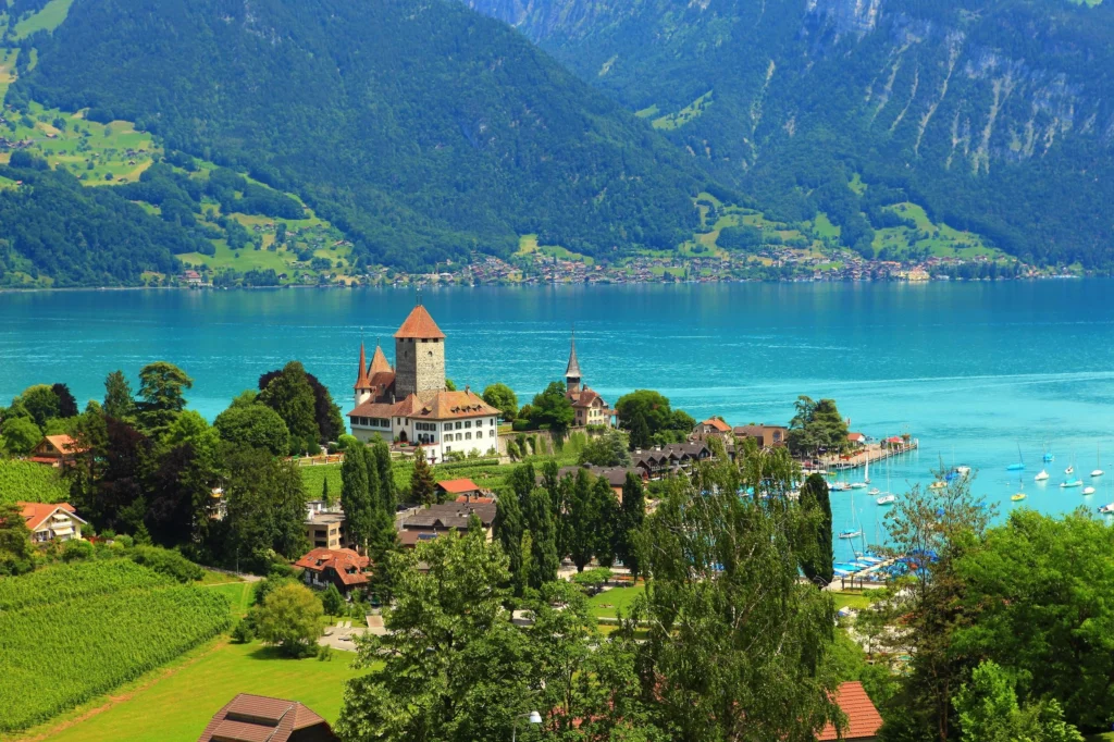 A beautiful view of the historic Spiez Castle situated on the shores of Lake Thun in Switzerland, with surrounding vineyards and mountain backdrop.