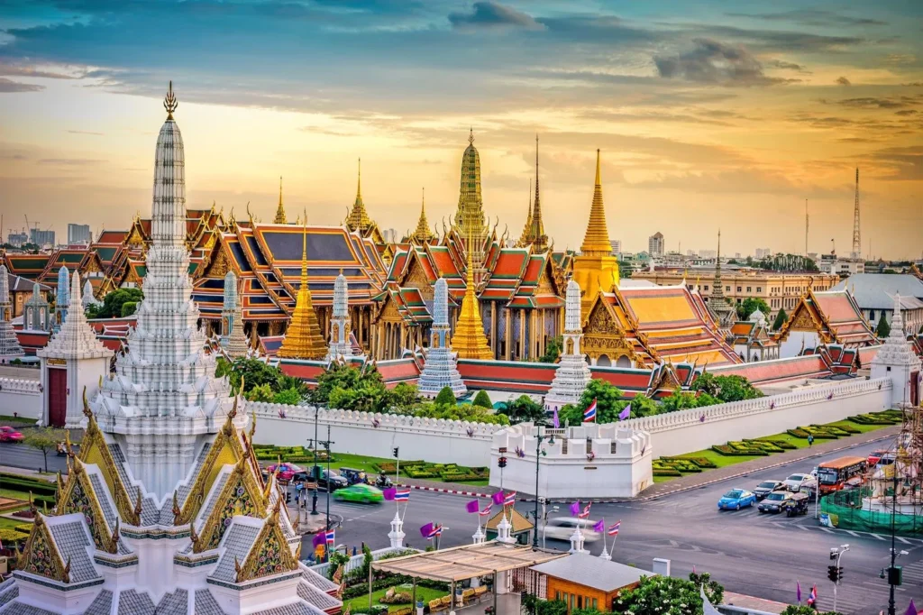 The stunning Grand Palace complex in Bangkok, Thailand, features ornate golden spires and colorful traditional roofs, glowing under a golden sunset.