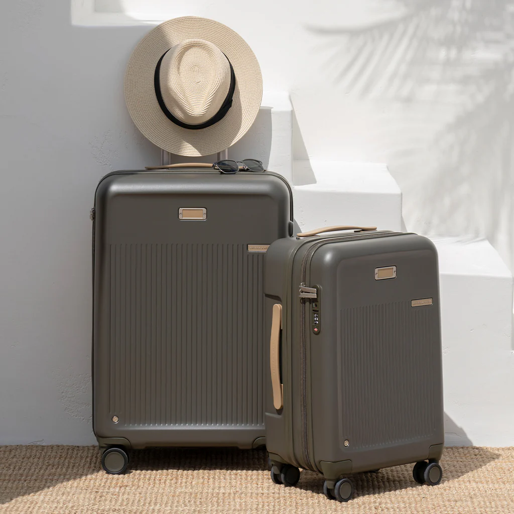 A set of two olive green hard-shell suitcases with a straw hat and sunglasses resting on them, set against a bright, minimalist backdrop.