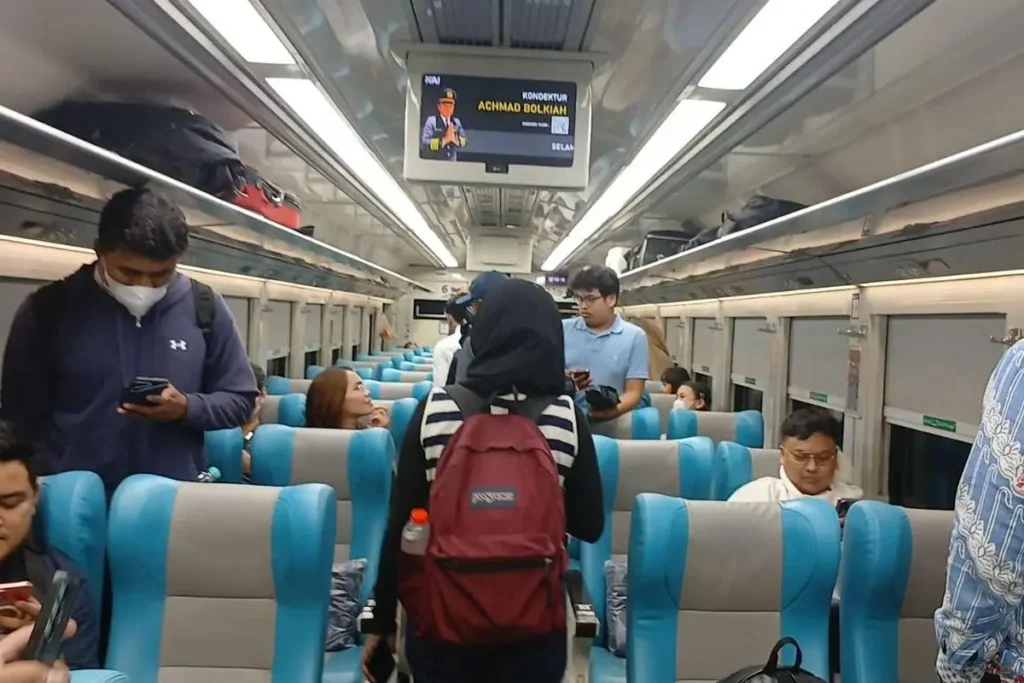 An Indonesian passenger train traveling along a track through lush green fields with a large majestic volcano rising in the background at sunrise.