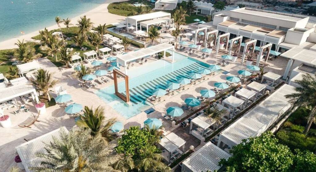 A high-angle view of a luxury beach club in Dubai, featuring a large swimming pool with blue umbrellas, cabanas, and a pristine sandy beach.