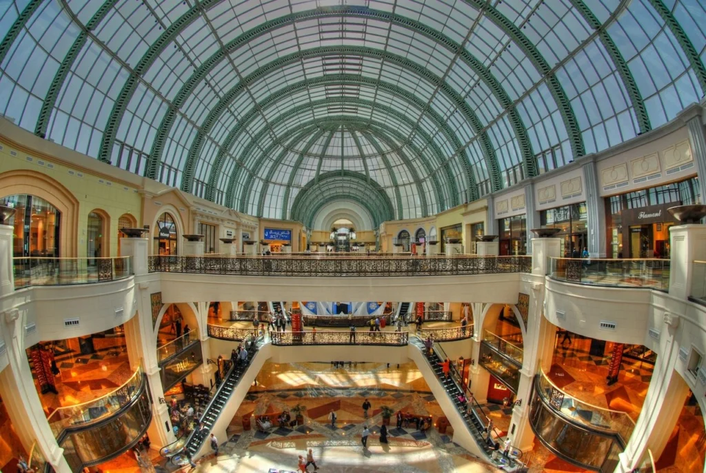 A fish-eye view of the multi-level interior of the Mall of the Emirates in Dubai, featuring a grand glass-domed ceiling, escalators, and shops.