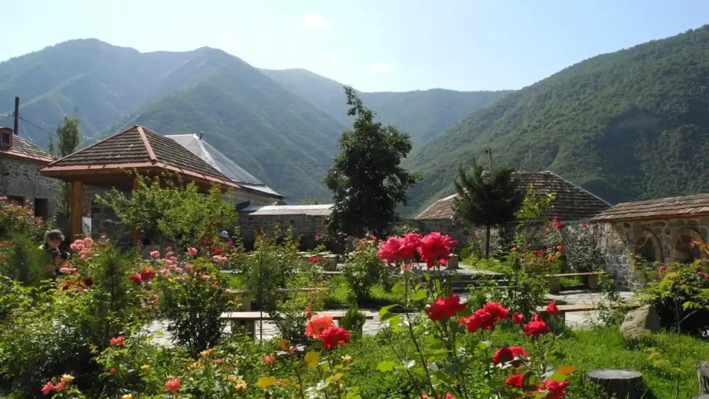 A view of the historic village of Kish near Sheki, Azerbaijan, featuring ancient stone architecture, cobblestone paths, and the iconic Kish Church nestled against a backdrop of lush, rolling green mountains.
