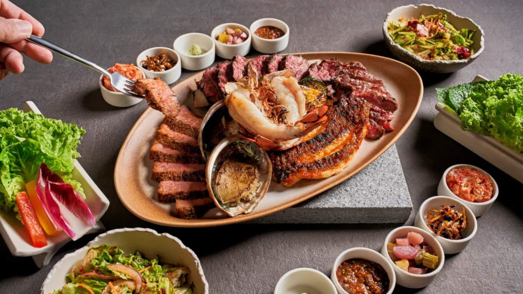 A premium platter in a Singapore restaurant, featuring grilled steak, lobster, and abalone, served with various side dishes and greens.