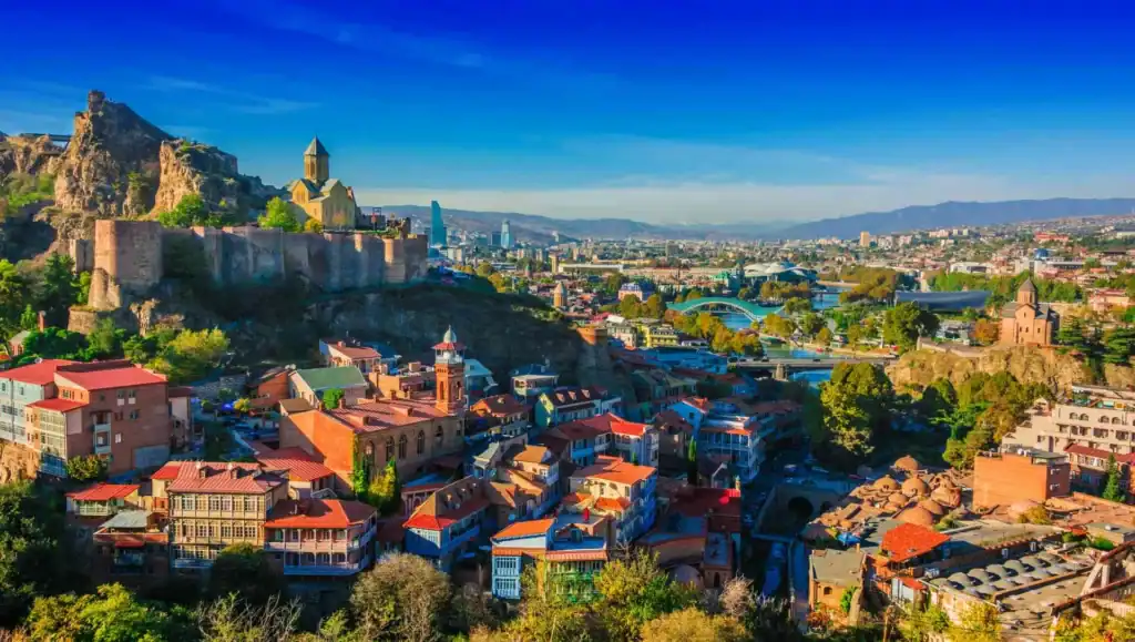 A picturesque cityscape of Tbilisi, Georgia, showcasing the colorful traditional houses with wooden balconies, the historic Narikala Fortress on the hill, and the modern Peace Bridge spanning the Mtkvari River.