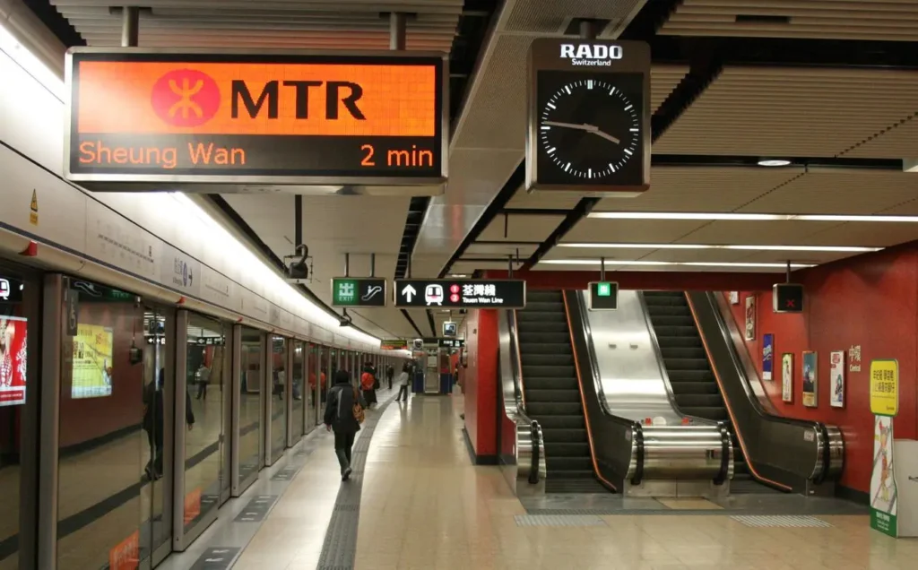 Interior view of the Sheung Wan MTR station platform in Hong Kong with digital train arrival signs, platform screen doors, and an escalator.