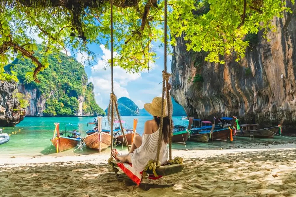 A woman in a sun hat sits on a wooden swing under a tree, looking out at traditional long-tail boats in a scenic tropical bay with limestone cliffs.
