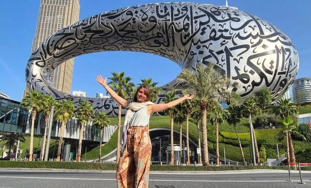 A woman standing with her arms outstretched in front of the iconic Museum of the Future building in Dubai, surrounded by palm trees.