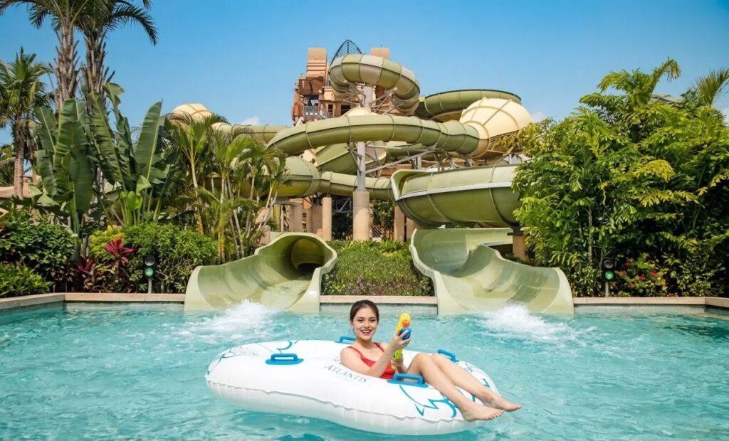 A woman smiling while floating on an inflatable ring in a water park pool, with large green water slides in the background.