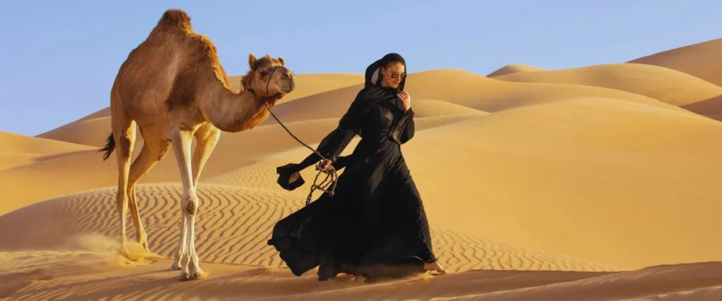 A woman in a black abaya and headscarf walking through sand dunes in the desert, accompanied by a camel on a lead.