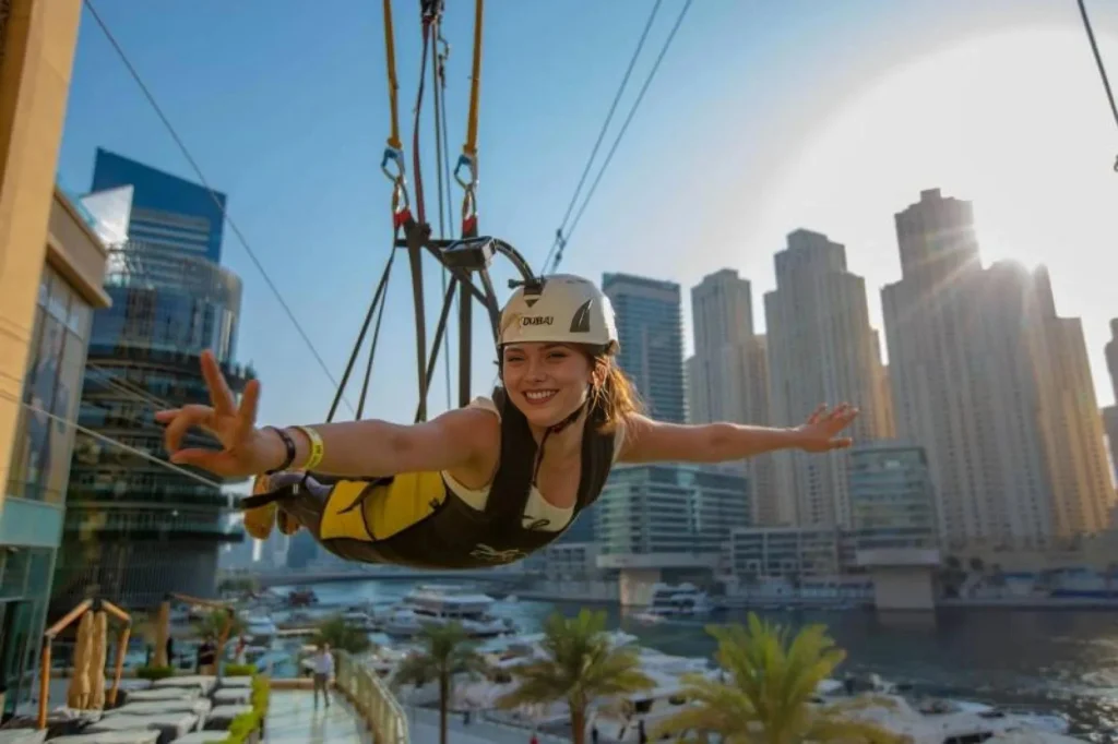 A smiling woman enjoying a zipline ride over the Dubai Marina, with the city's iconic skyscrapers and waterfront in the background.