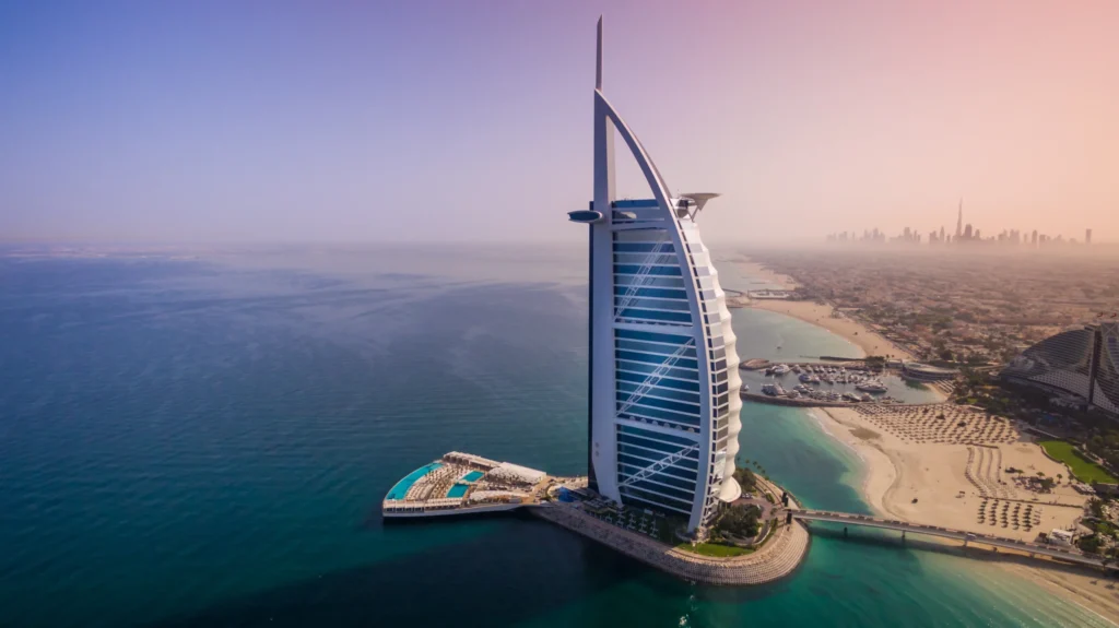 An aerial view of the iconic sail-shaped Burj Al Arab hotel in Dubai, situated on its own island surrounded by clear blue waters and a sandy beach.