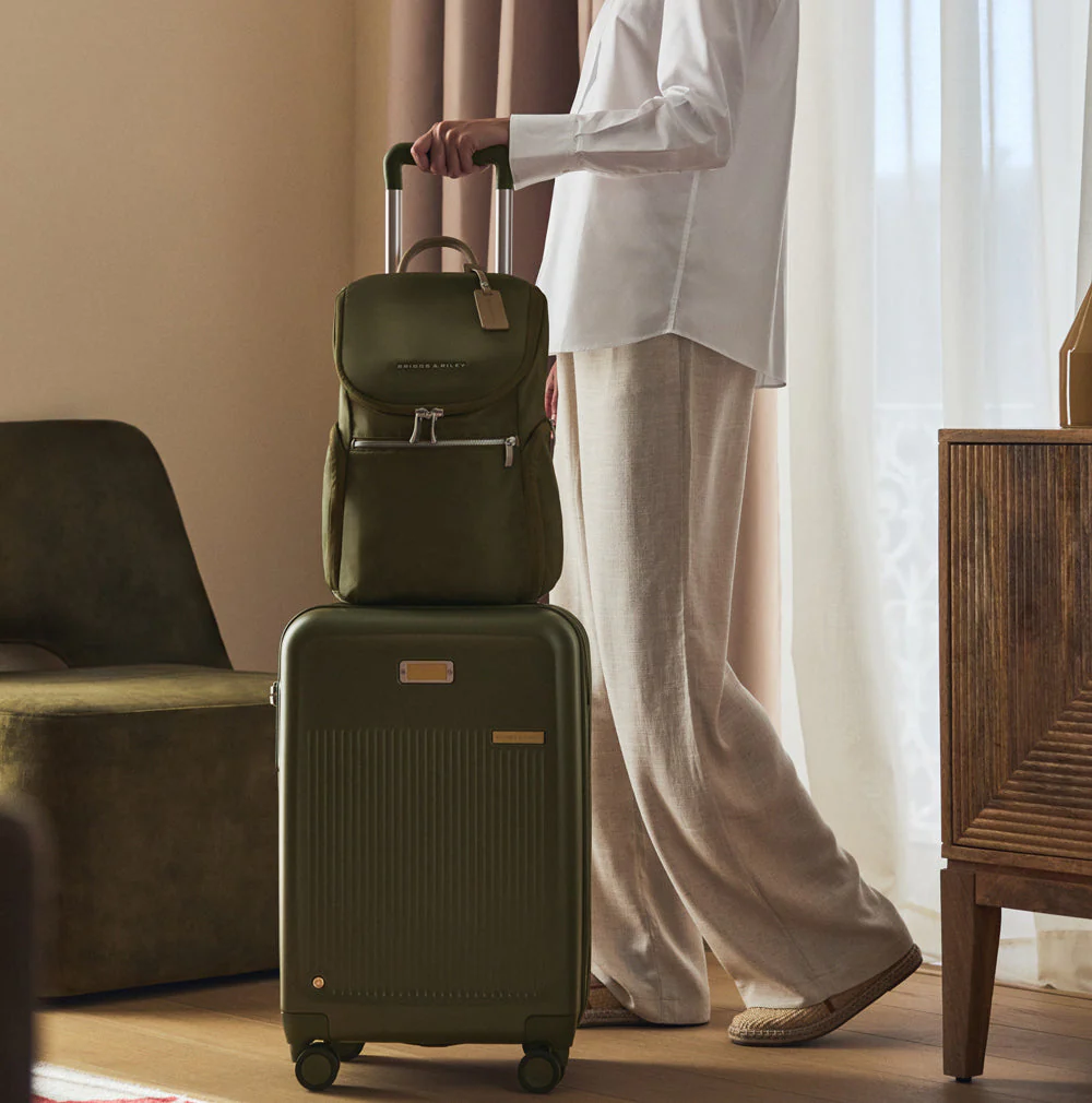 A set of two olive green hard-shell suitcases with a straw hat and sunglasses resting on them, set against a bright, minimalist backdrop.