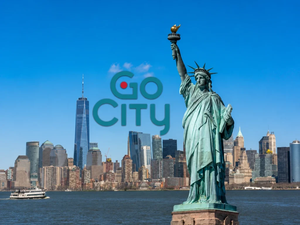 The Statue of Liberty holding a torch stands on the right, with the New York city skyline and One World Trade Center in the background under a clear blue sky, featuring the Go City logo.