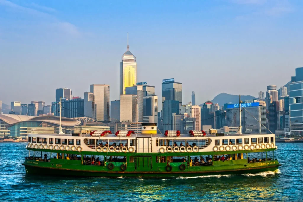 A traditional green and white Star Ferry cruising across Victoria Harbour with the Hong Kong Island skyline and skyscrapers in the background.