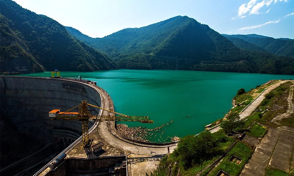 A wide view of the massive concrete arch of the Enguri Dam in Svaneti, Georgia, set against a backdrop of deep green mountains and clear water.