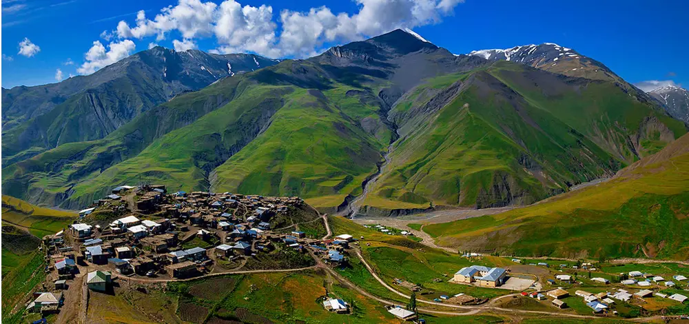 An aerial view of the historic Khinalug village nestled on a hilltop, surrounded by lush green slopes and the towering peaks of the Caucasus Mountains in Azerbaijan.
