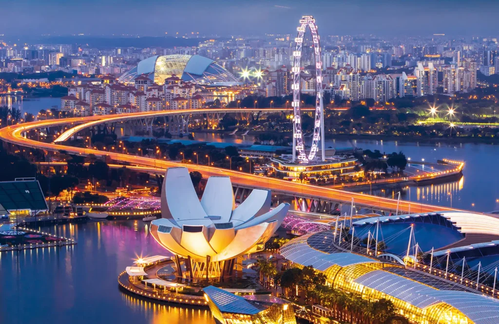 An illuminated night view of Singapore featuring the ArtScience Museum, the Singapore Flyer, and vibrant city lights reflecting on the water.