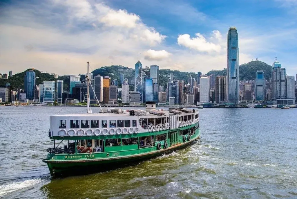 A traditional green and white Star Ferry cruising across Victoria Harbour with the Hong Kong Island skyline and skyscrapers in the background.