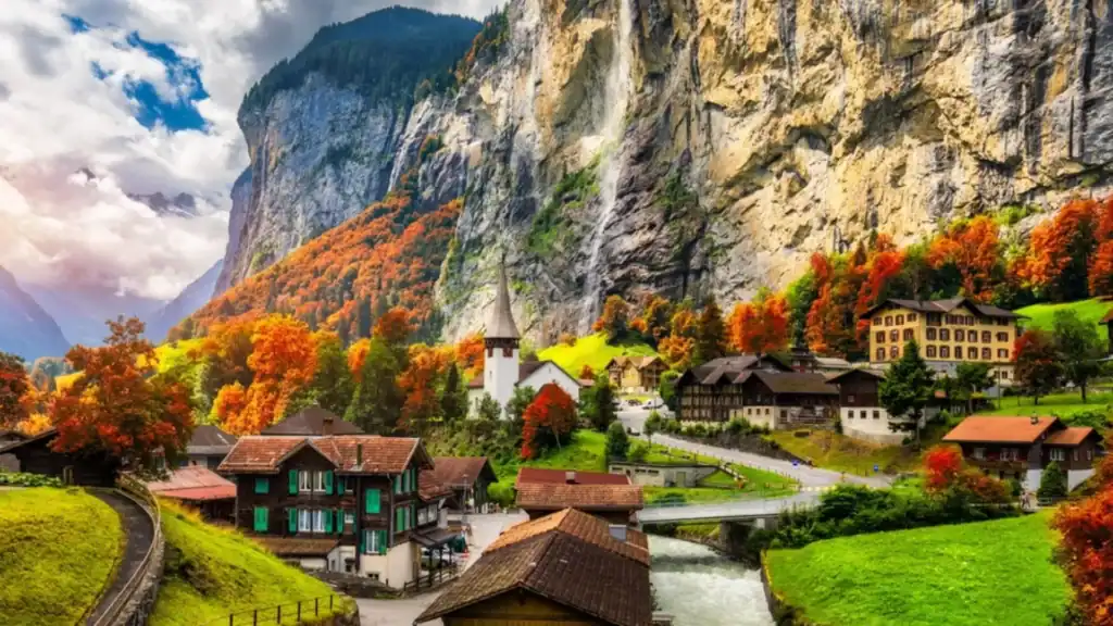 A breathtaking panoramic view of the Lauterbrunnen valley in Switzerland, featuring towering limestone cliffs, lush green meadows, a picturesque village, and the iconic Staubbach Falls cascading in the distance.