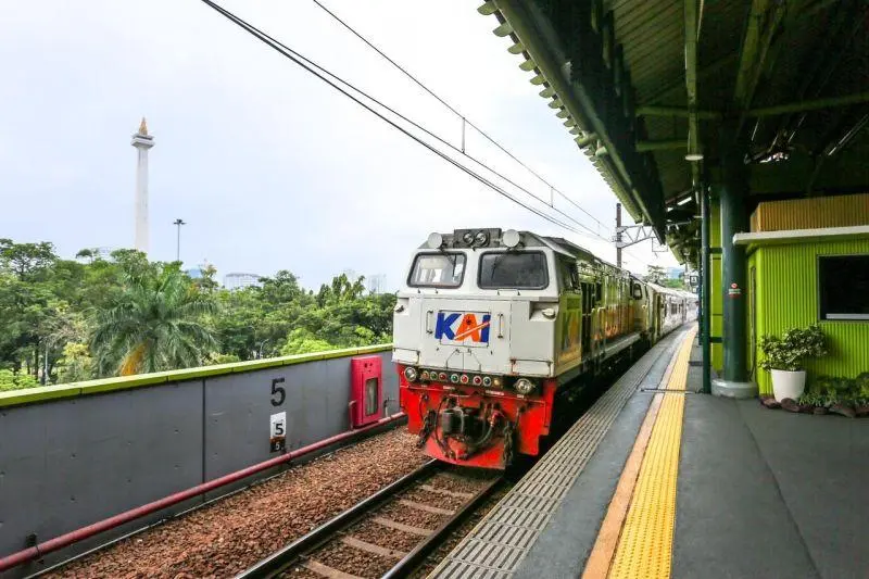 An Indonesian passenger train traveling along a track through lush green fields with a large majestic volcano rising in the background at sunrise.