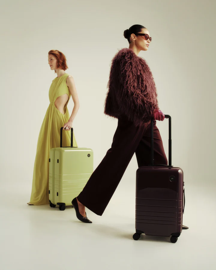Two fashionably dressed women walking with matching light green and deep mulberry-colored hard-shell suitcases against a plain backdrop.