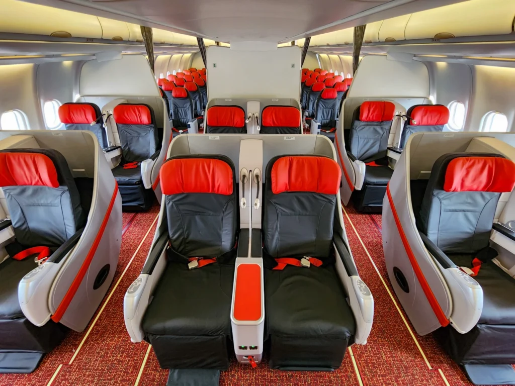 A symmetrical interior view of an airplane business class cabin featuring comfortable seats with red and black leather headrests and a red carpet aisle.
