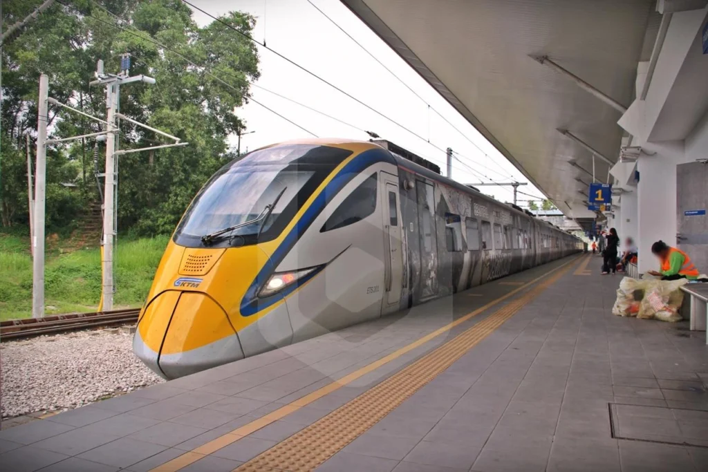 A high-speed KTM ETS electric train with a yellow and blue nose parked at a concrete station platform with passengers waiting in the background.