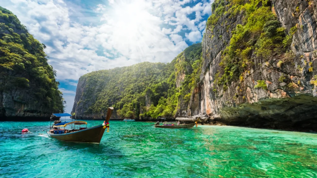 A traditional Thai long-tail boat floats on crystal-clear turquoise water in a sunny cove surrounded by lush, towering limestone cliffs.