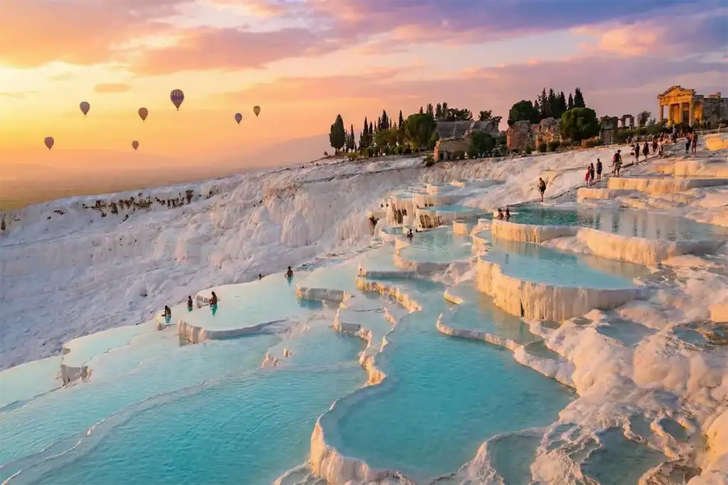 Scenic sunset view of Pamukkale, Turkey, featuring bright white travertine terraces filled with turquoise thermal pools and hot air balloons above.