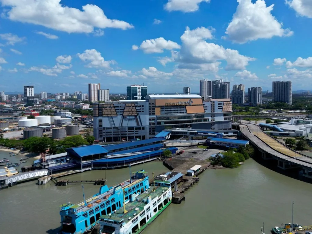An aerial view of Penang Sentral transportation hub with parked ferries in the foreground under a bright blue sky with fluffy white clouds.