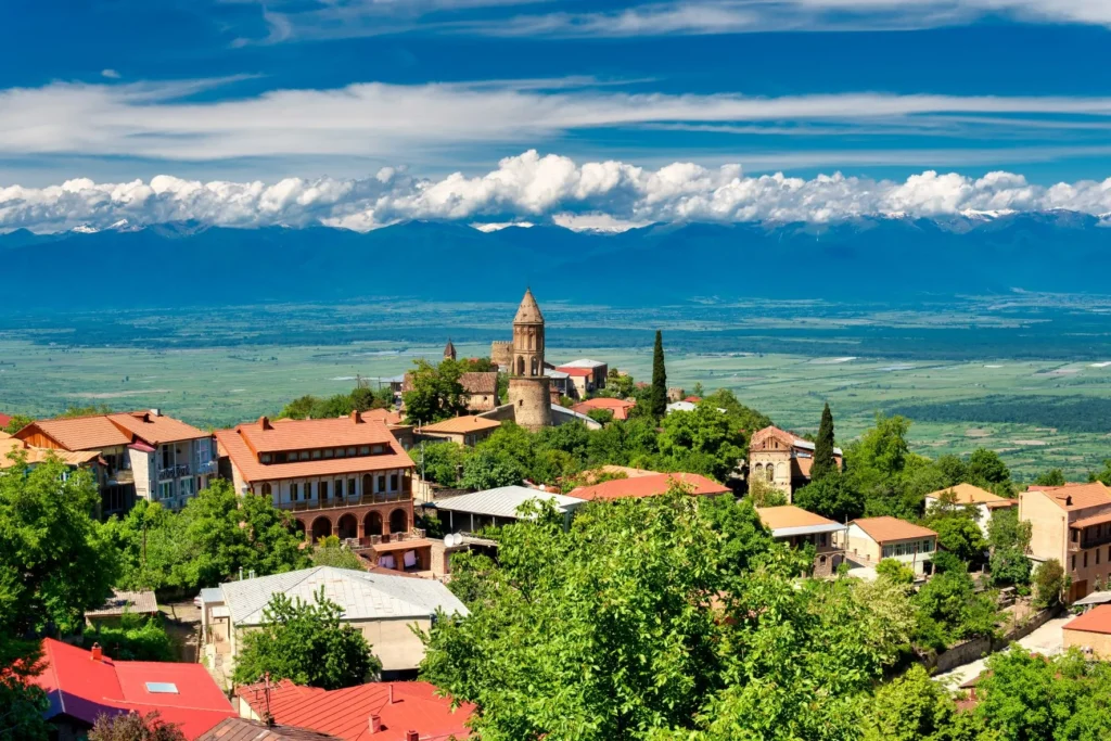 A scenic view of the hillside town of Sighnaghi in Georgia, featuring red-roofed buildings, ancient stone walls, and vast mountain landscapes.
