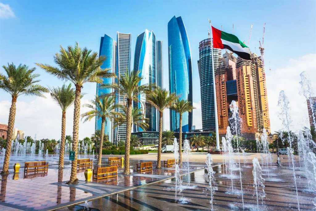The Abu Dhabi skyline with iconic blue glass skyscrapers and the UAE flag flying, set behind a public plaza with palm trees and water fountains.