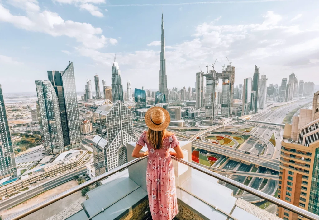A tourist with a hat and backpack waving at the iconic Dubai Marina skyscrapers and a traditional wooden dhow boat on the water.