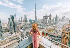 A tourist with a hat and backpack waving at the iconic Dubai Marina skyscrapers and a traditional wooden dhow boat on the water.