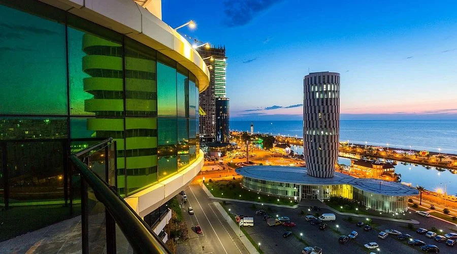 A panoramic city view from a hotel balcony, overlooking dense urban rooftops, streets, and distant buildings under a bright daylight sky.