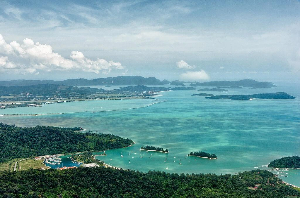 An aerial landscape view of the lush green coastline of Langkawi, featuring a small marina, turquoise waters, and distant islands under a blue sky.