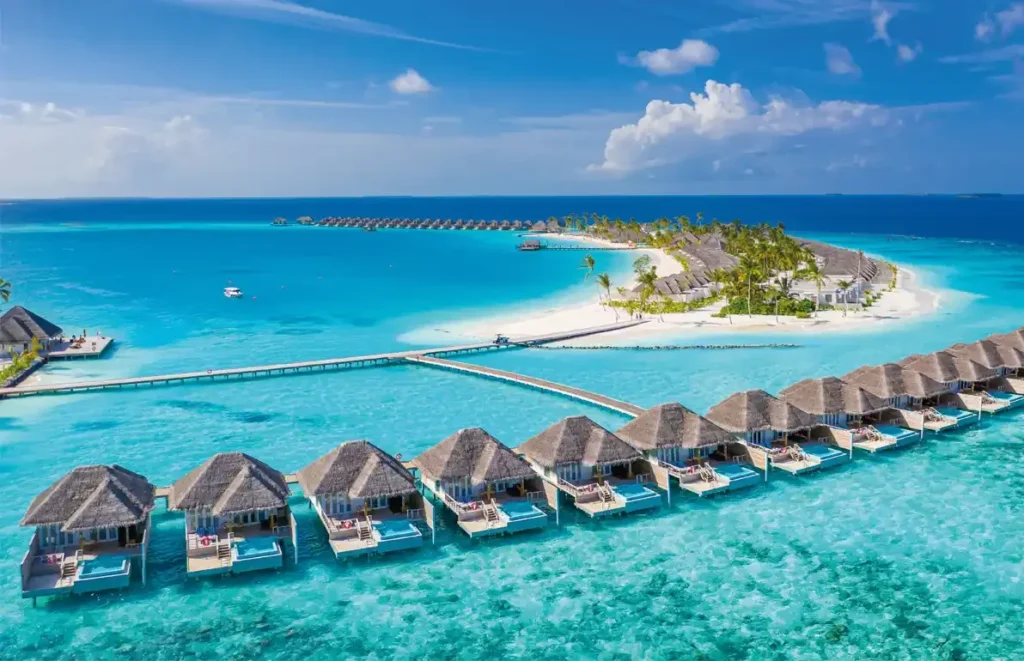 A wooden walkway leads to a restaurant built over turquoise water at a tropical island resort, surrounded by palm trees and a blue sky.